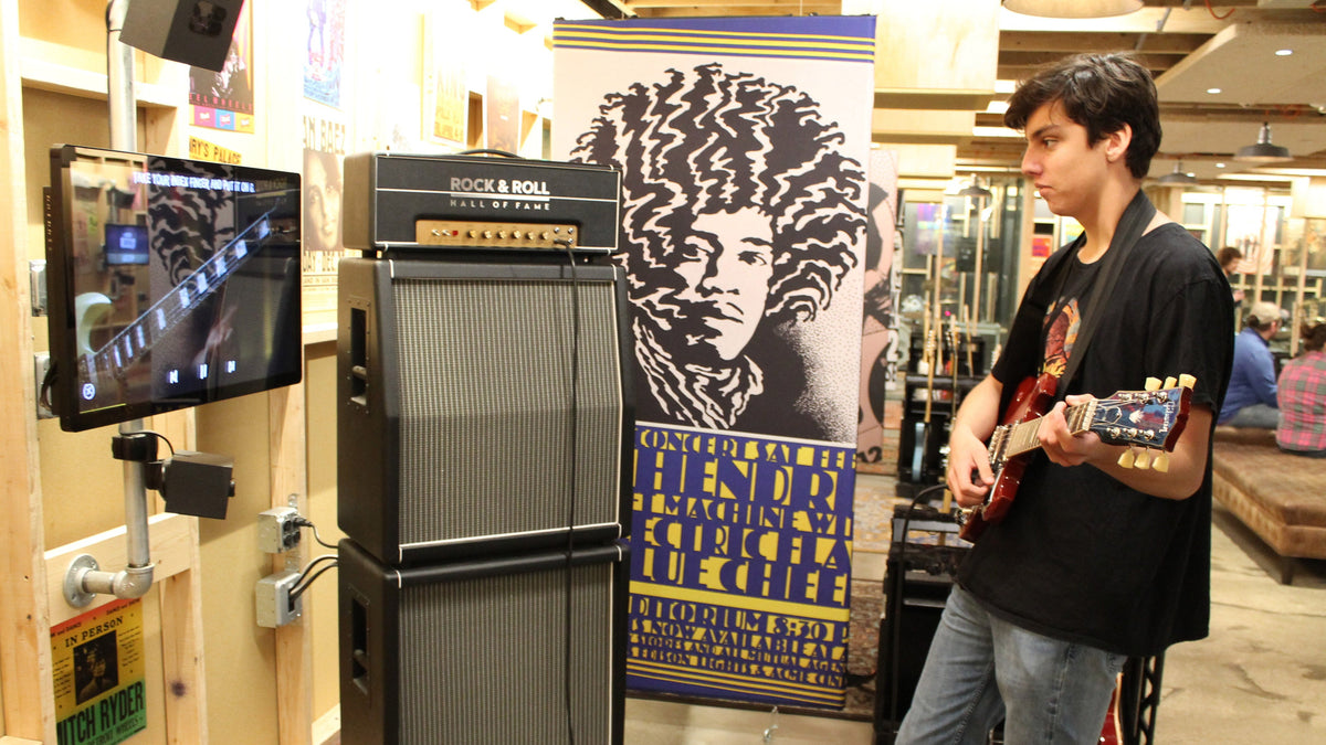 A teenager playing electric guitar at a Garage learn-to-play station, with a touchscreen on an arm beside the amp stack